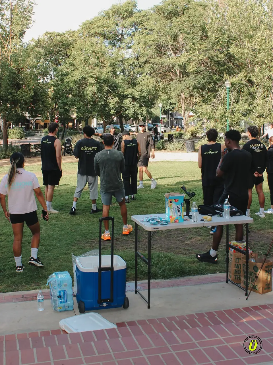 Crew gathering on the grass at Shelton Park with a cooler and snack table nearby