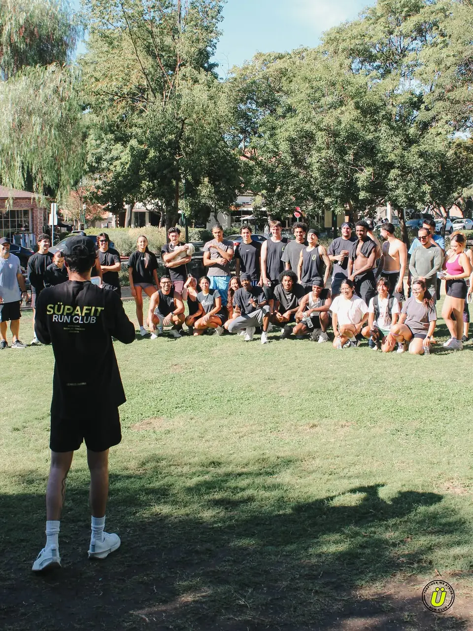Host in a SÜPAFIT Run Club shirt talking to the whole crew on the grass
